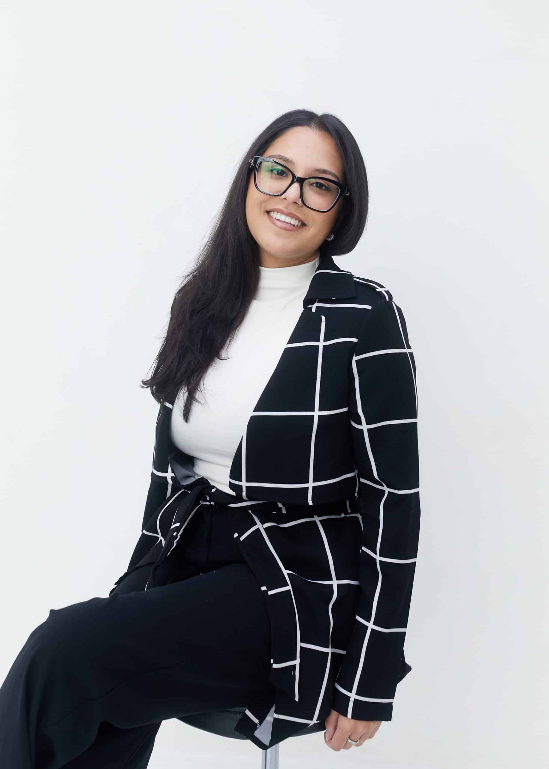 Photo of clinic manager of VIP MEDSPA - Osanna Ramkisson - sitting on a chair wearing black pants and a white top with a black and white jacket, smiling. 
